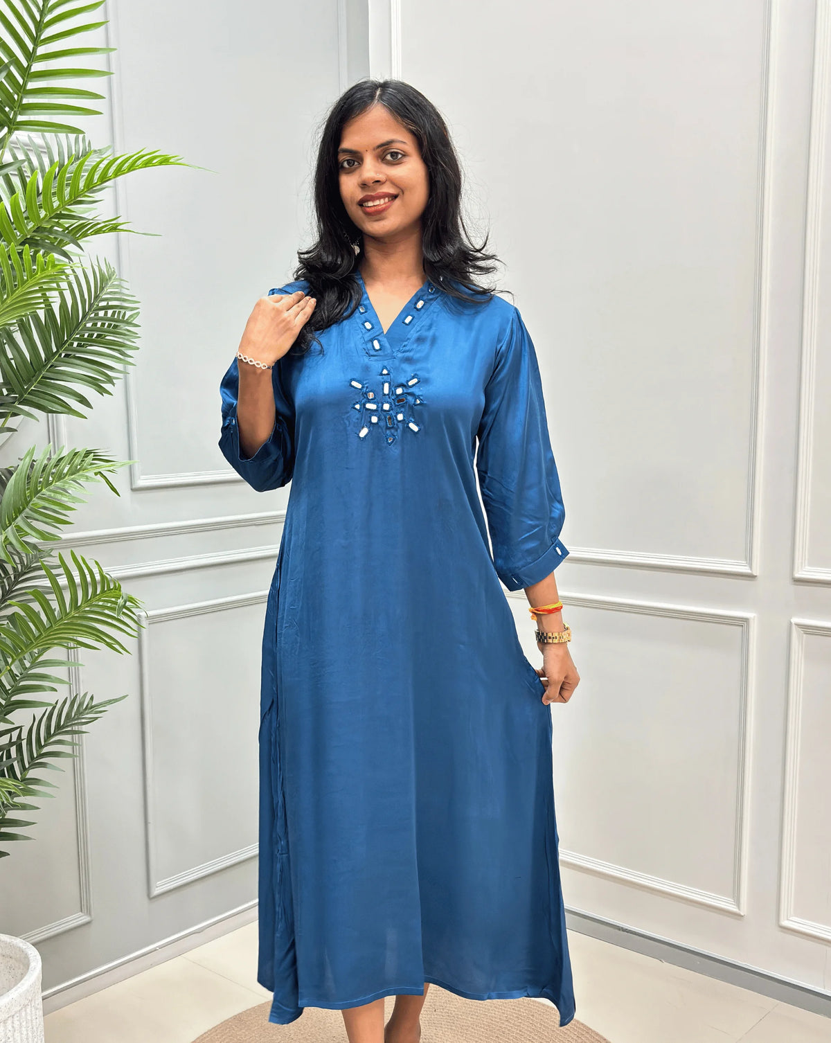 Front -View of Blue Women's Pure Modal Silk Ajrakh Long Kurti with a decorative Real Hand-Mirror embroidered V-neckline, Floral pattern on front & Sleeves Cuffs, standing on a round woven rug in front of a white paneled wall with a potted plant on the left