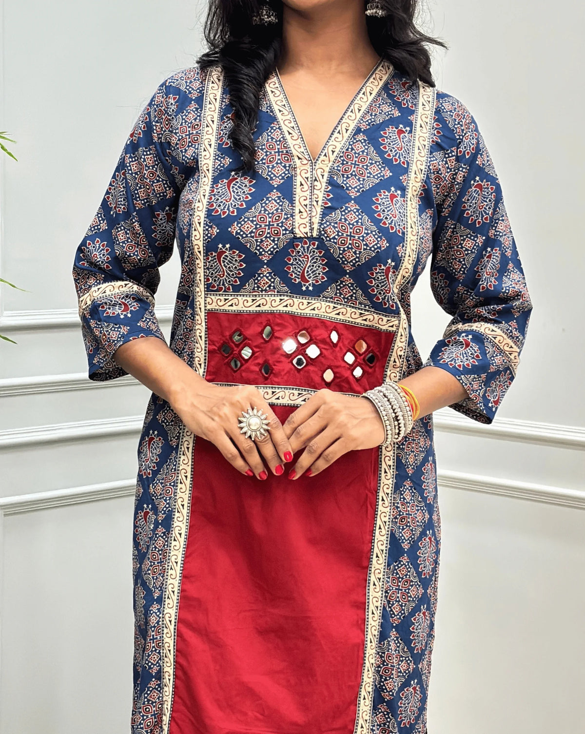 Close-up of Red Women's Ajrakh pure cotton 2 peice Co-Ord set featured with V Shaped neck with a Blue base & an Peacock print on Front Chest to Side & Sleeves, Hand-mirror work on Front as Belt, standing on a round woven mat in front of a white paneled wall with a potted plant.