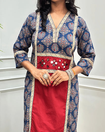 Close-up of Red Women's Ajrakh pure cotton 2 peice Co-Ord set featured with V Shaped neck with a Blue base & an Peacock print on Front Chest to Side & Sleeves, Hand-mirror work on Front as Belt, standing on a round woven mat in front of a white paneled wall with a potted plant.