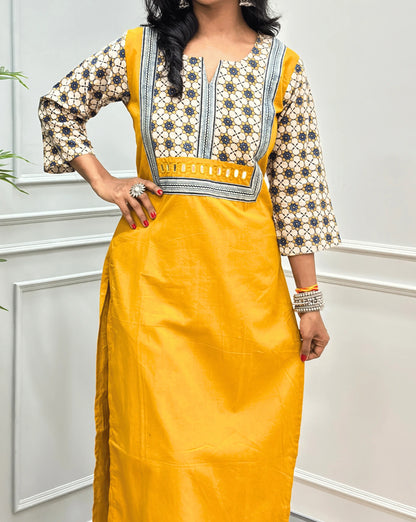 Close-up view of Mustard Yellow Women's Hand-block Ajrakh Pure Cotton Long Kurti featured with Round V Shaped neck along with three quarter sleeves, with a Beige base , an abstract of Blue & Black Floral Print yoke on front & Full sleeves, Hand-mirror work on Front, paired with blue Pant, standing on a round woven mat in front of a white paneled wall.