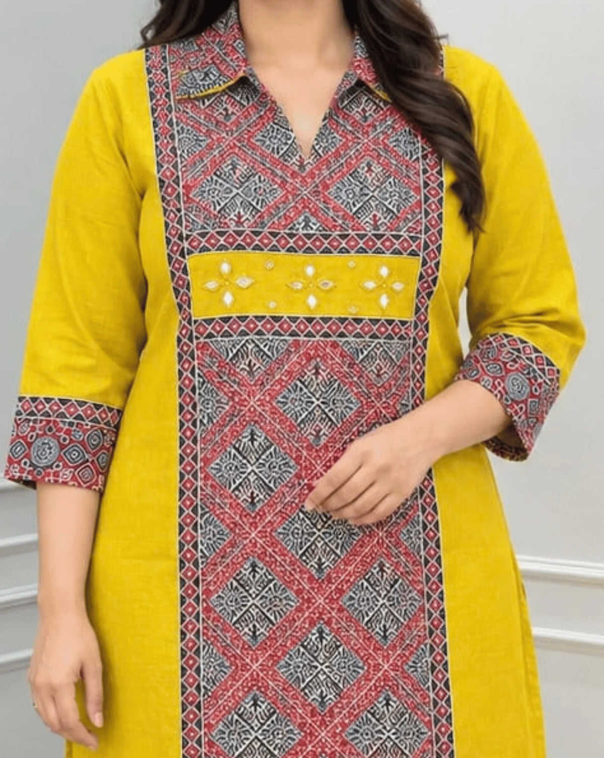 Close-up of Mustard Yellow Ajrakh Women's  Vegetable Dyed Plant Based Cotton Long-Kurti Featured with Full pattren Collar V shaped Neck along with Three quarter Sleeves, with a Red Base,  an Abstract of Black & Beige geometric patterned panel on front & Sleeves Cuffs, Real Hand-Mirror Patch work on Front, paired with Red pants, standing in a bright room with a white & Grey paneled wall, on left side a potted plant.