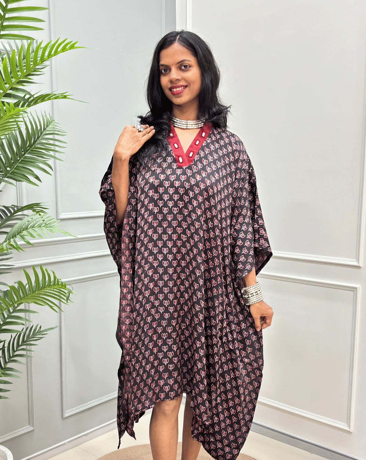 Front-view of Black Women's Ajrakh Pure Modal Silk Knee Length Kaftan with a black Floral print, with Contrast red Colour V-neck, standing on a round woven rug in front of a white paneled wall next to a potted plant. 