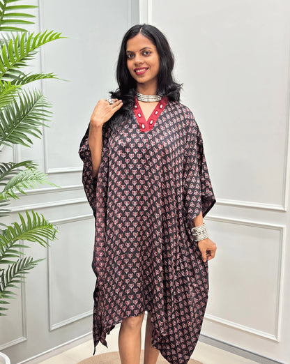 Front-view of Black Women's Ajrakh Pure Modal Silk Knee Length Kaftan with a black Floral print, with Contrast red Colour V-neck, standing on a round woven rug in front of a white paneled wall next to a potted plant. 