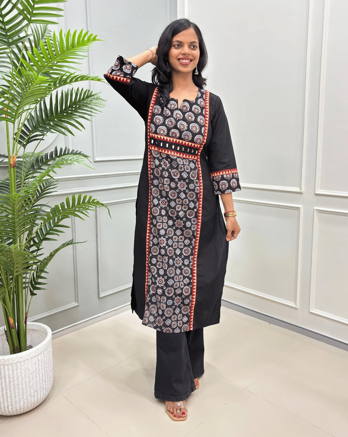 Front view of Black Ajrakh hand-block long-kurti with blue, red and white floral print, red & beige border outline detailed , paired with black wide-leg pants, standing in a bright room with a potted plant.