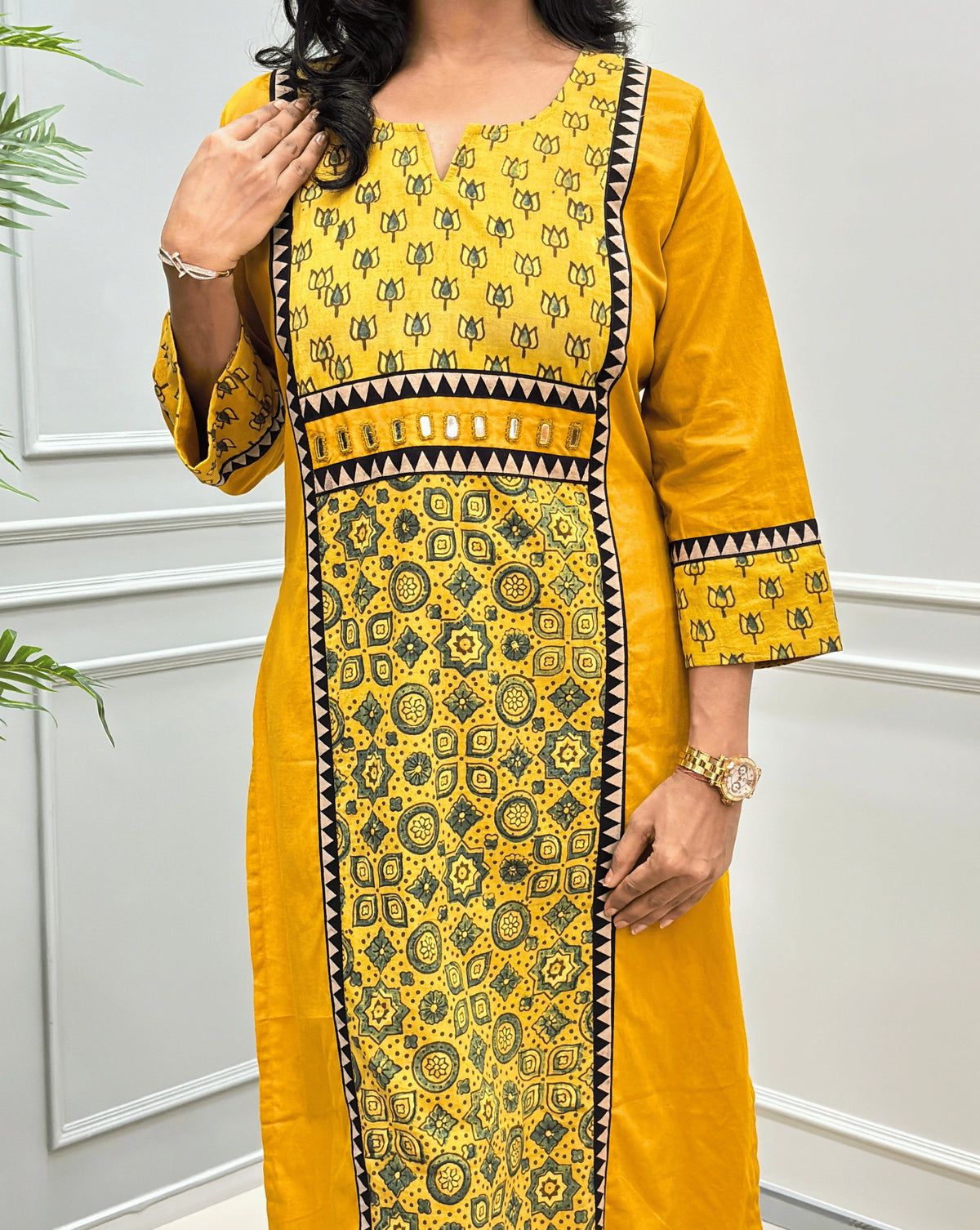 Close-up of mustard yellow Ajrakh long-kurti with yellow and green geometric patterns, shown from the neck to down in panel, Hand-mirror work on waist , standing against a white paneled wall.