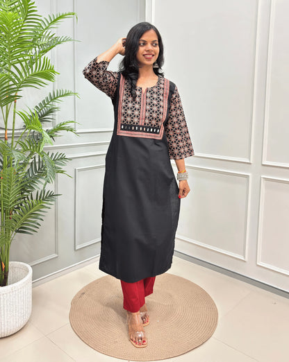 Black Women's Hand-block Ajrakh Pure Cotton Long Kurti with a Black base , an abstract of Red & beige Floral Print yoke on front & Full sleeves, paired with blue Pant, standing on a round woven mat in front of a white paneled wall.
