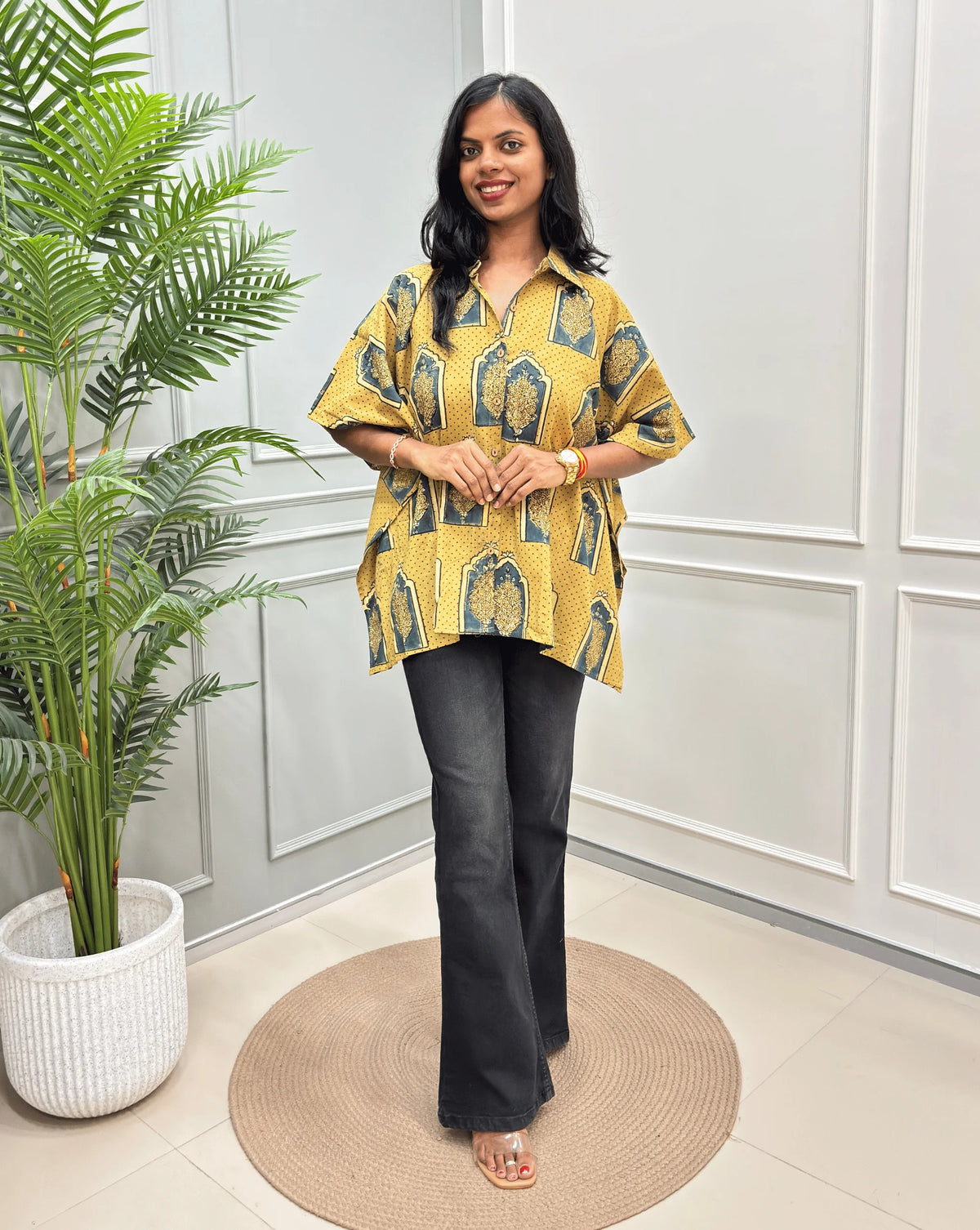 Light Yellow Women's Ajrakh Pure Cotton Short Kaftan Shirt with a Beige, Green & Yellow Floral Block Print, paired with beige flared pants, standing on a round woven rug in front of a white paneled wall with a potted plant.