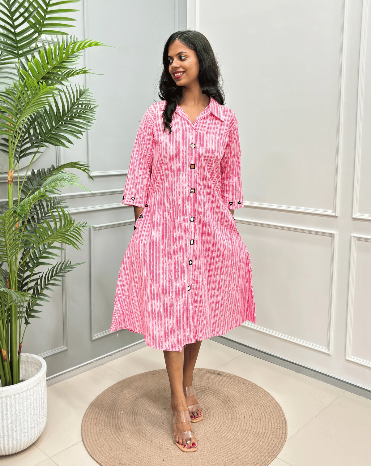 Pink Women's Ajrakh A-line Pure Cotton Long Kurti with an Ajrakh Strip Print on abstract of Pink, White standing on a round woven rug in front of a light gray paneled wall with a potted plant.