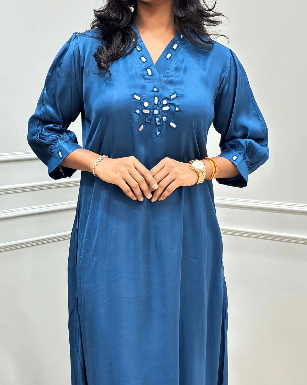 Close-up view of Blue Women's Pure Modal Silk Ajrakh Long Kurti Featured with 3/4th Sleeves with a decorative Real Hand-Mirror embroidered V-neckline, Floral pattern on front & Sleeves Cuffs, standing on a round woven rug in front of a white paneled wall with a potted plant on the left