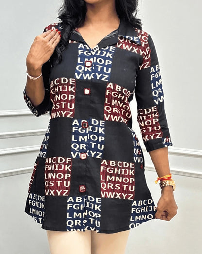 Close up of Black Women's Ajrakh Vegetable Dyed Plant based Cotton Short kurti featured with Collar neck & three Quarter sleeves with a Red & Blue Base, & Abstract Beige color alphabet print, Hand-mirror work on Front, Sleeves & Pockets,  paired with beige trousers, standing on a round woven rug in front of a white paneled wall with a potted plant.