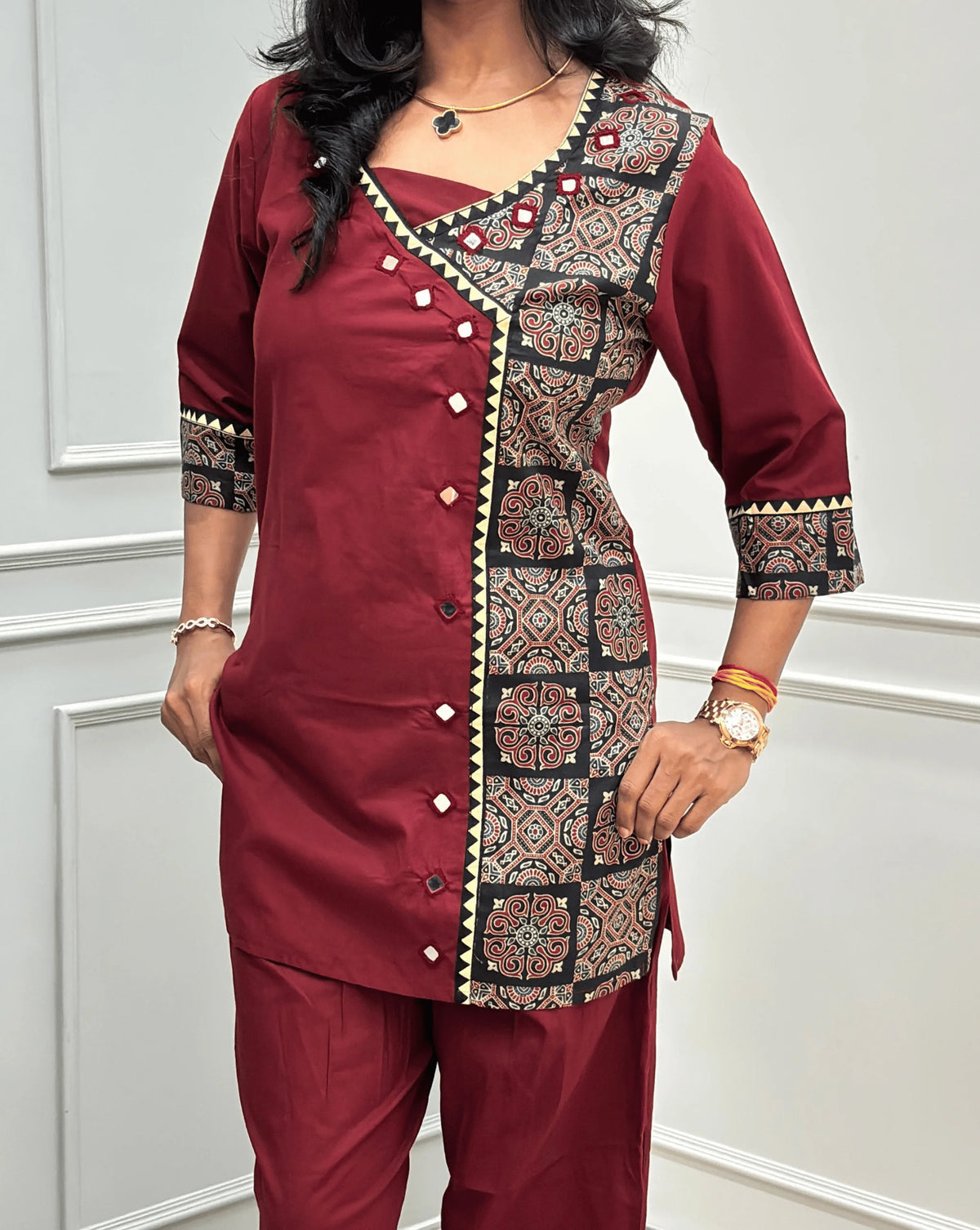 Close-up view of Red Women's Ajrakh Pure Cotton 2-peice Co-Ord set Featured with V-shaped neck with a Ajrakh printed panel on the front & Sleeves, Angrakha Patterned on front, Real Hand-Mirror work on front Along with 2 side pockets in pant, standing on a round woven rug in a modern room with white paneled walls.
