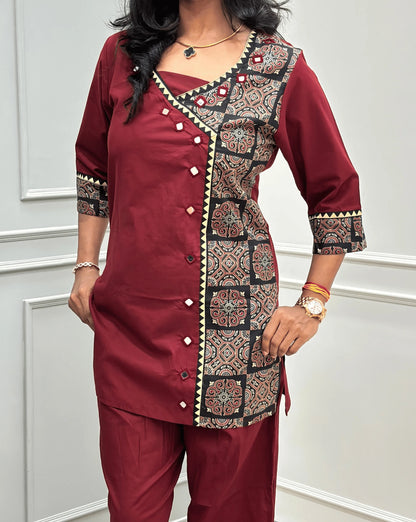 Close-up view of Red Women's Ajrakh Pure Cotton 2-peice Co-Ord set Featured with V-shaped neck with a Ajrakh printed panel on the front & Sleeves, Angrakha Patterned on front, Real Hand-Mirror work on front Along with 2 side pockets in pant, standing on a round woven rug in a modern room with white paneled walls.