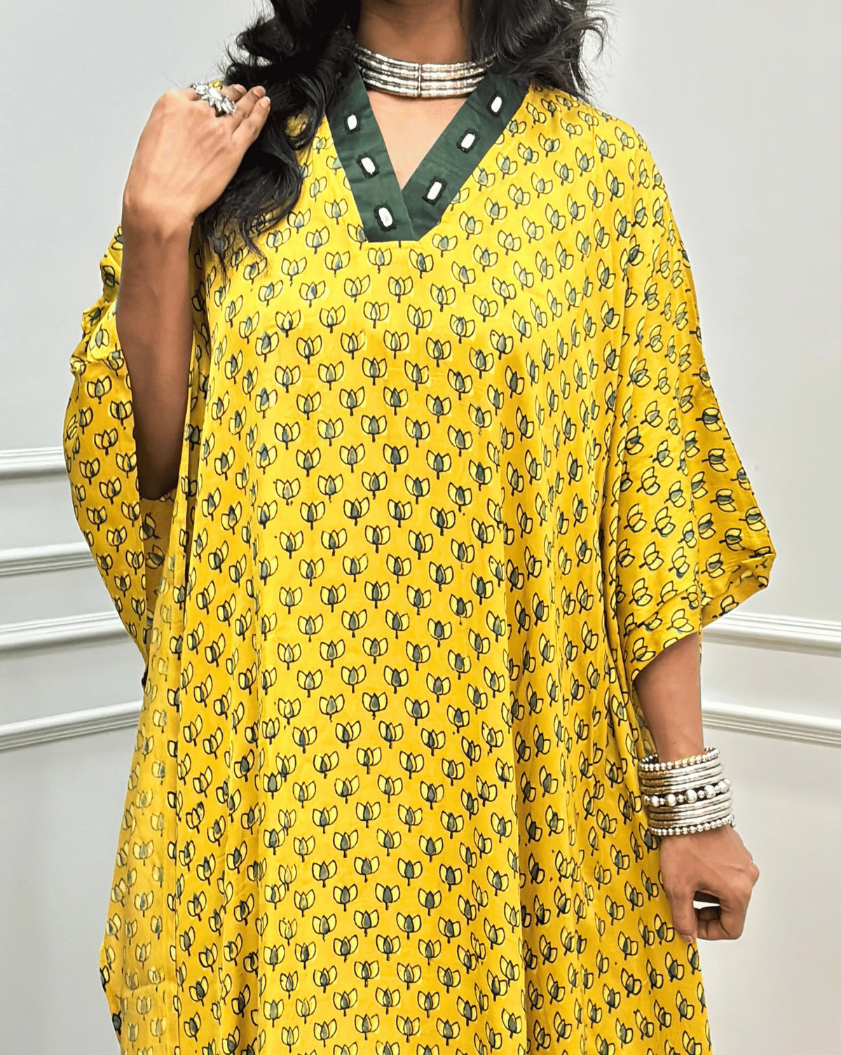 Close-up view of Yellow Women's Ajrakh Pure Modal Silk Knee Length Kaftan with a black Floral print, with Contrast Green Colour V-neck, along with Real Hand-Mirror embroidery, standing on a round woven rug in front of a white paneled wall next to a potted plant. 