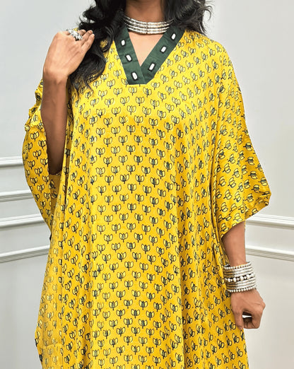Close-up view of Yellow Women's Ajrakh Pure Modal Silk Knee Length Kaftan with a black Floral print, with Contrast Green Colour V-neck, along with Real Hand-Mirror embroidery, standing on a round woven rug in front of a white paneled wall next to a potted plant. 