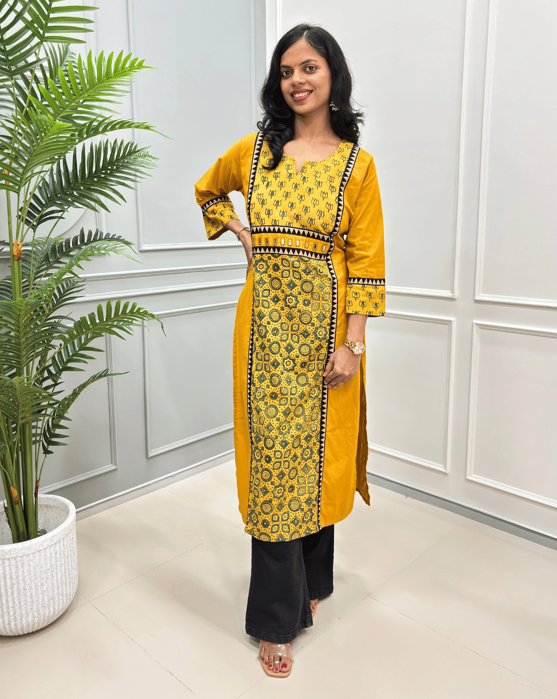 Mustard Yellow Women's cotton long-  kurti with yellow and green geometric patterns, Hand-mirror work on waist like a belt black-yellow border detailed on  front and sleeves with paired with black wide-leg pants, standing in an indoor setting with a potted plant on the left.