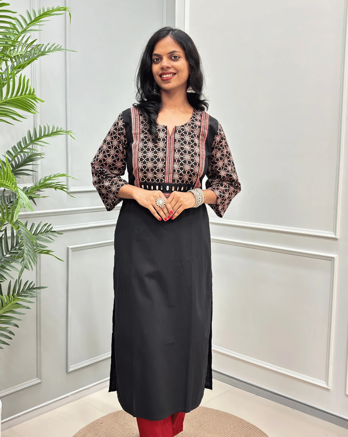 Front view of Black Women's Hand-block Ajrakh Pure Cotton Long Kurti with a Black base , an abstract of Red & beige Floral Print yoke on front & Full sleeves, Hand-mirror work  on front,  paired with blue Pant, standing on a round woven mat in front of a white paneled wall.