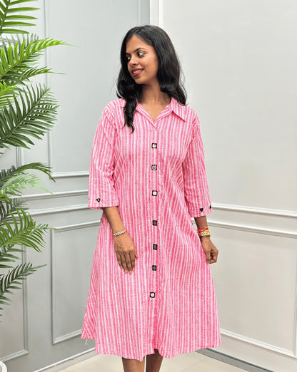 Front-View of Pink Women's Ajrakh A-line Pure Cotton Long Kurti with an Ajrakh Strip Print on abstract of Pink, White,  Real hand-mirror work on front & Sleeves, Along with 2 side pockets, standing on a round woven rug in front of a light gray paneled wall with a potted plant.