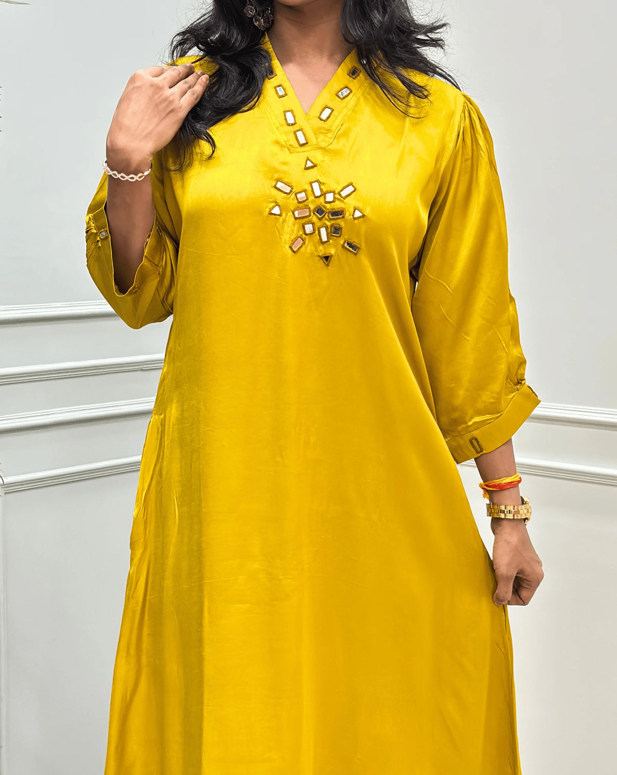 Close-up view of Yellow Women's Pure Modal Silk Ajrakh Long Kurti Feature with 3/4 th Sleeves with a decorative Real Hand-Mirror embroidered V-neckline, Floral pattern on front & Sleeves Cuffs, standing on a round woven rug in front of a white paneled wall with a potted plant on the left
