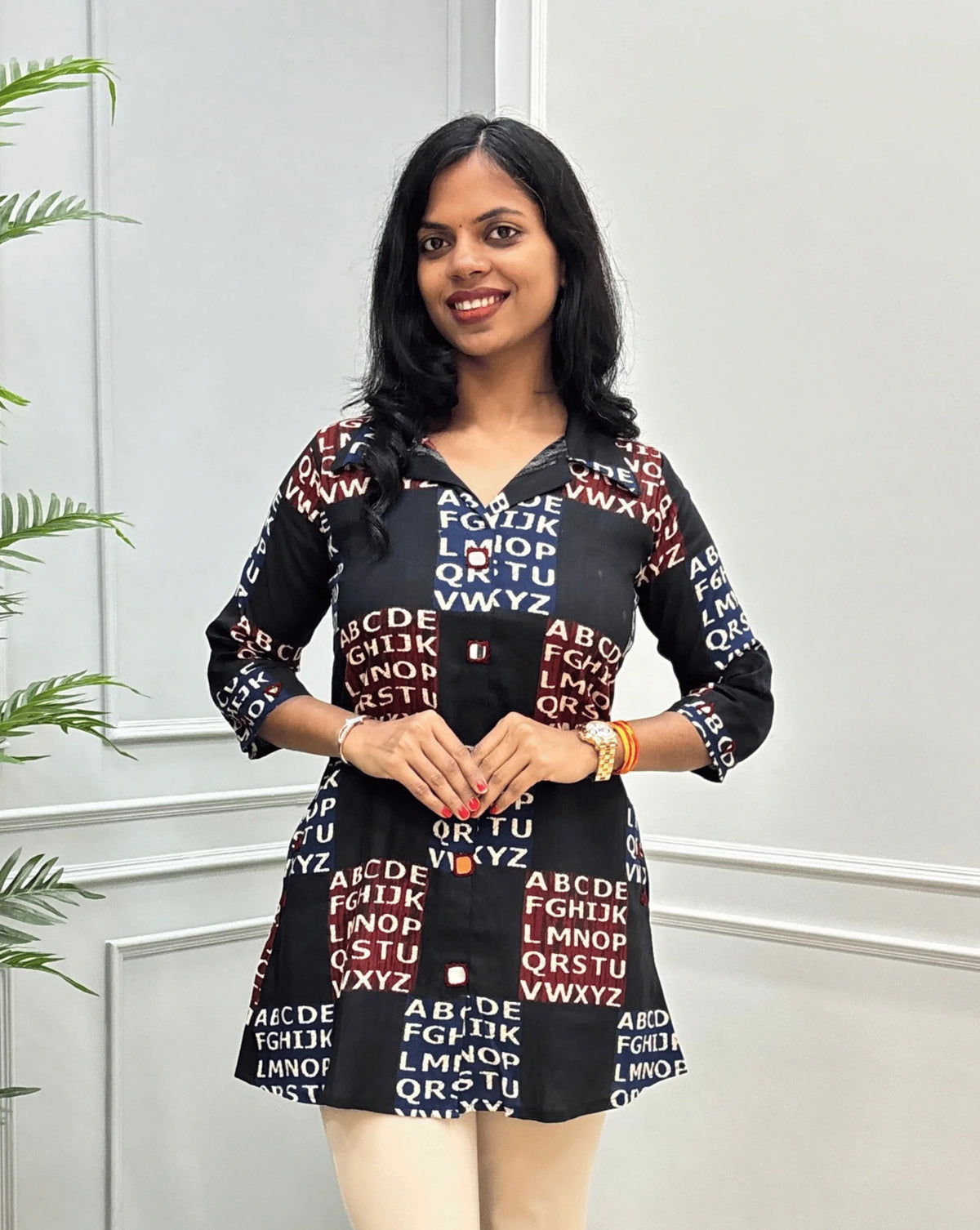 Front view of Black Women's Ajrakh Vegetable Dyed Plant based Cotton Short kurti with a Red & Blue Base, & Abstract Beige color alphabet print, Hand-mirror work on Front, Sleeves & Pockets,  paired with beige trousers, standing on a round woven rug in front of a white paneled wall with a potted plant.