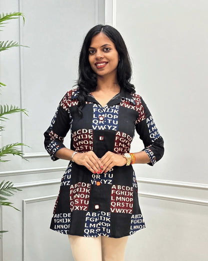 Front view of Black Women's Ajrakh Vegetable Dyed Plant based Cotton Short kurti with a Red & Blue Base, & Abstract Beige color alphabet print, Hand-mirror work on Front, Sleeves & Pockets,  paired with beige trousers, standing on a round woven rug in front of a white paneled wall with a potted plant.