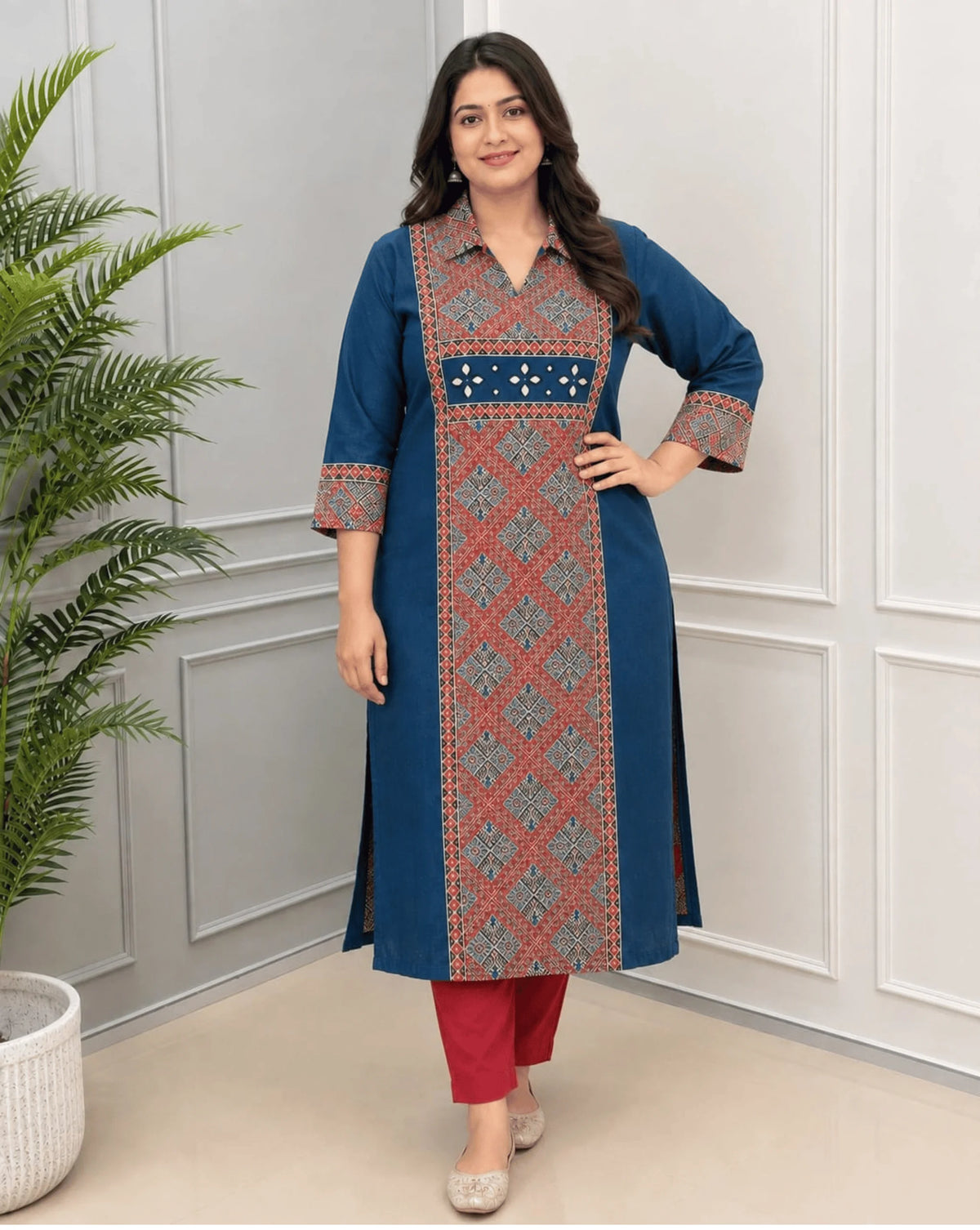 Deep Blue Women's Ajrakh Vegetable Dyed Plant Based Cotton Long-Kurti with a red Base,  an Abstract of Black  & Beige geometric patterned panel on front & Sleeves Cuffs, paired with red pants, standing in a bright room with a white & Grey paneled wall ,on left side a potted plant.