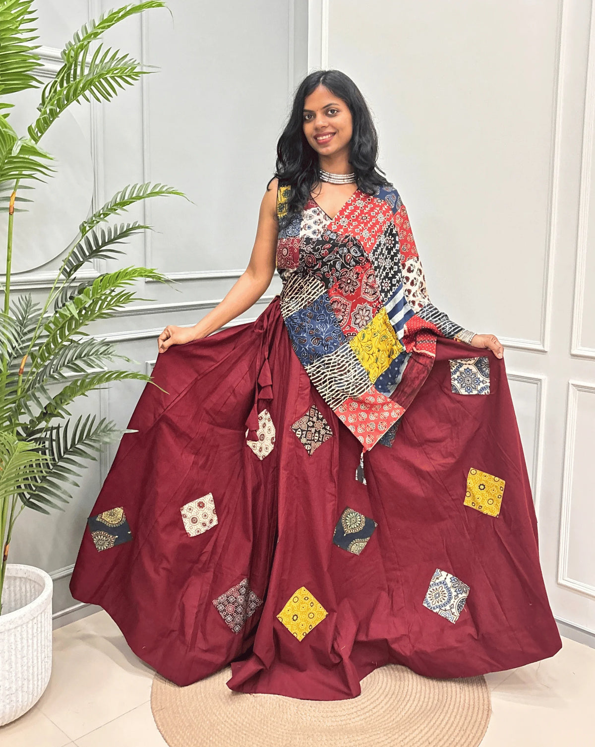 Red Women's Ajrakh pure Cotton Skirt Set with a beautiful Tasals & Ajrakh Patch work along with multi-colour Printed blouse & Dupatta, Standing against a light paneled wall with a green plant on the left.
