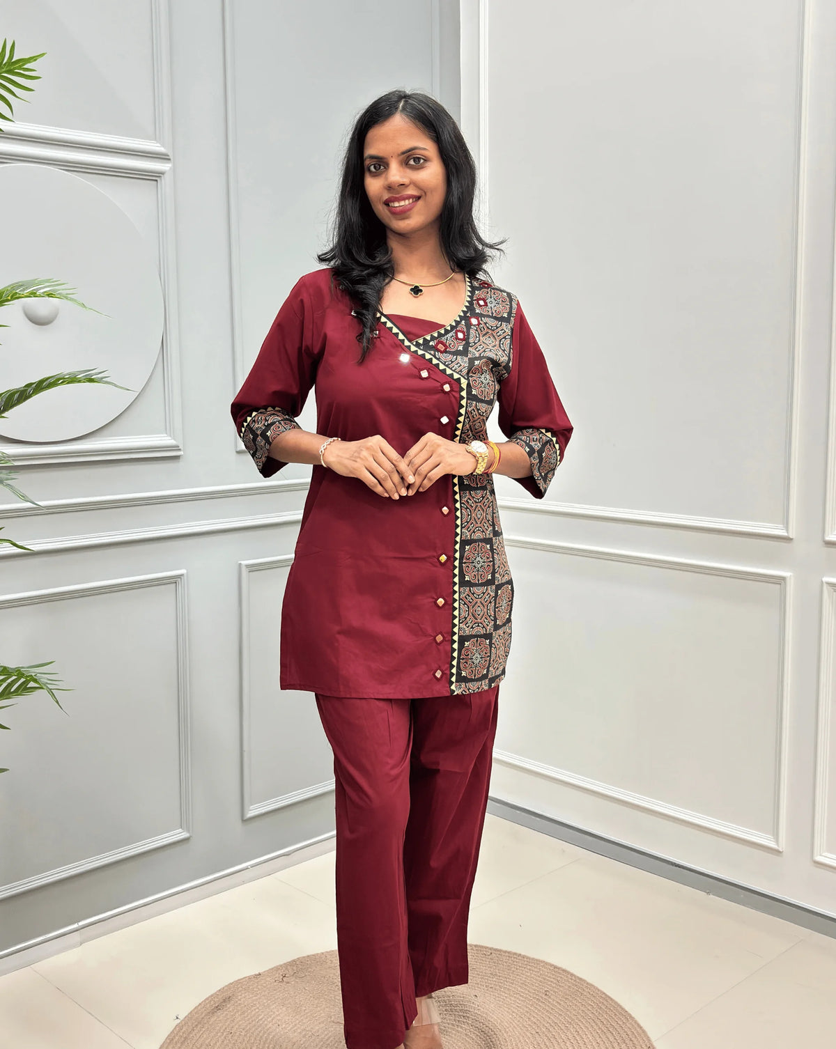 Front-view of Red Women's Ajrakh Pure Cotton 2-peice Co-Ord set with a Ajrakh printed panel on the front & Sleeves, Angrakha Patterned on front, Real Hand-Mirror work on front Along with 2 side pockets in pant, standing on a round woven rug in a modern room with white paneled walls.