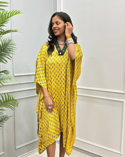 Front-view of Yellow Women's Ajrakh Pure Modal Silk Knee Length Kaftan with a black Floral print, with Contrast Green Colour V-neck, standing on a round woven rug in front of a white paneled wall next to a potted plant.