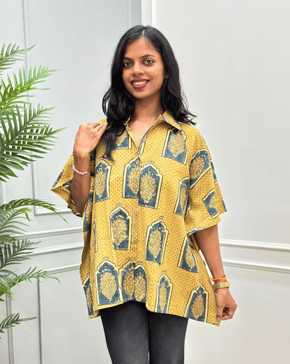 Front view of Light Yellow Women's Ajrakh Pure Cotton Short Kaftan Shirt with a Beige, Green & Yellow Floral Block Print, Wooden Button on front, paired with beige flared pants, standing on a round woven rug in front of a white paneled wall with a potted plant.