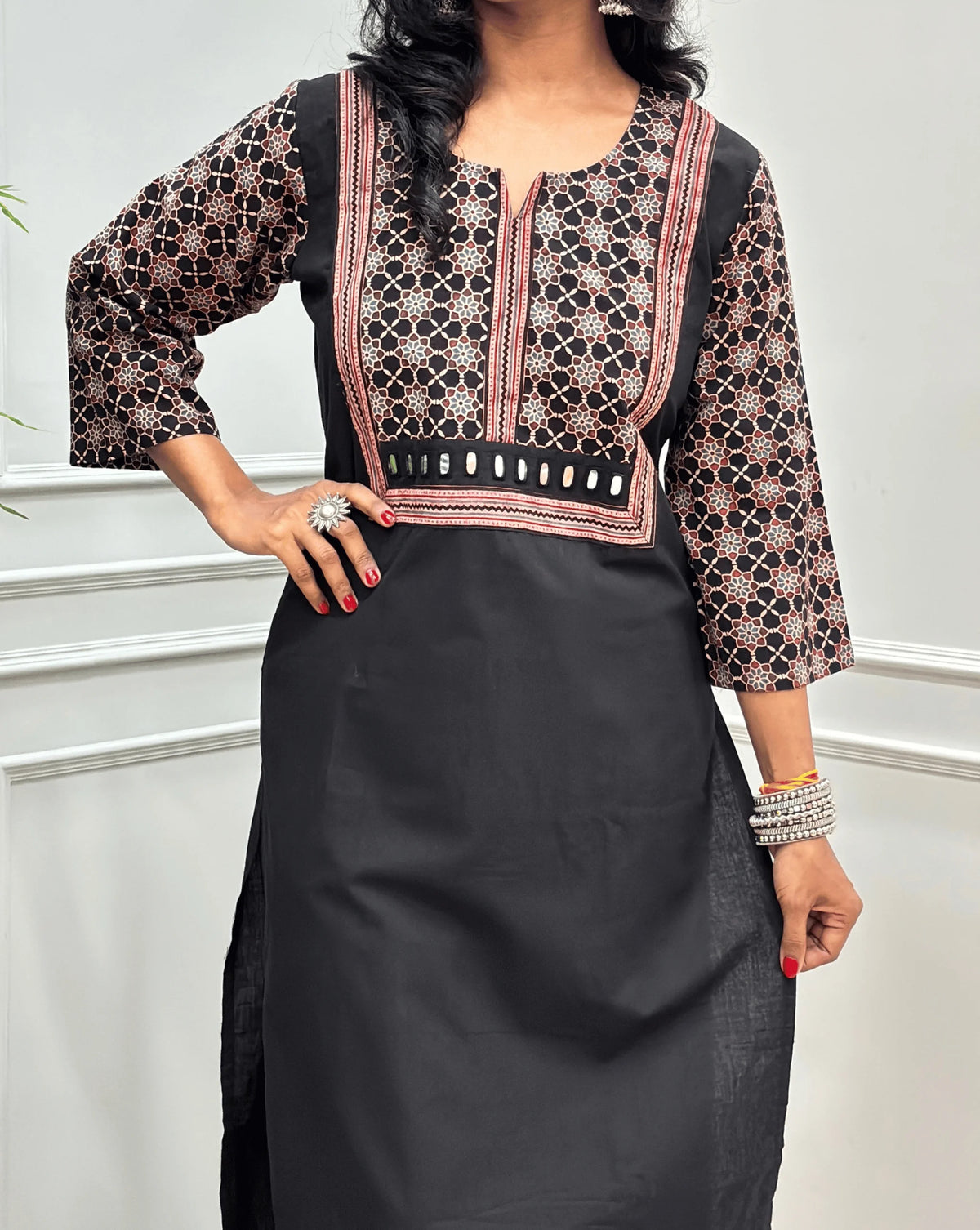 Close-up view of Black Women's Hand-block Ajrakh Pure Cotton Long Kurti featured with Round V Shaped neck along with three quarter sleeves, with a Black base , an abstract of Red & beige Floral Print yoke on front & Full sleeves, Hand-mirror work  on front,  paired with blue Pant, standing on a round woven mat in front of a white paneled wall.