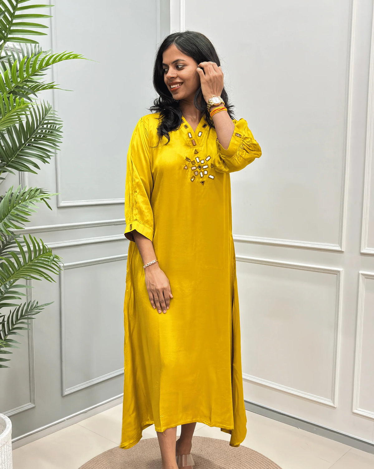 Front -View of Yellow Women's Pure Modal Silk Ajrakh Long Kurti with a decorative Real Hand-Mirror embroidered V-neckline, Floral pattern on front & Sleeves Cuffs, standing on a round woven rug in front of a white paneled wall with a potted plant on the left