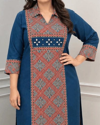 Close-up of Deep Blue Women's Ajrakh Vegetable Dyed Plant Based Cotton Long-Kurti Featured with Full pattren Collar V shaped Neck along with Three quarter Sleeves, with a Red Base,  an Abstract of Black & Beige geometric patterned panel on front & Sleeves Cuffs, Real Hand-Mirror Patch work on Front, paired with Red pants, standing in a bright room with a white & Grey paneled wall, on left side a potted plant.