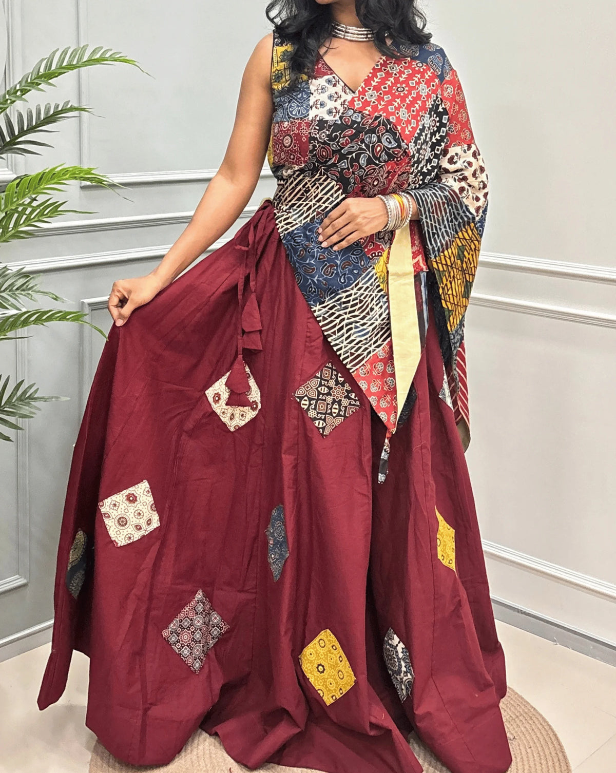 Close-up View of Red Women's Ajrakh pure Cotton Skirt Set with a beautiful Tasals & Ajrakh Patch work along with multi-colour Printed blouse & Dupatta, Standing against a light paneled wall with a green plant on the left.
