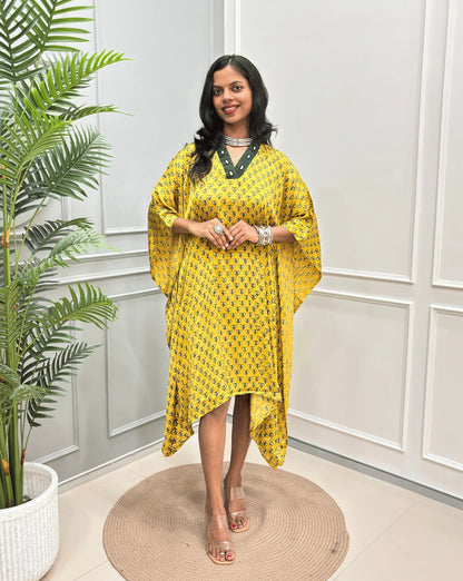 Yellow Women's Ajrakh Pure Modal Silk Knee Length Kaftan with a black Floral print , standing on a round woven rug in front of a white paneled wall next to a potted plant.
