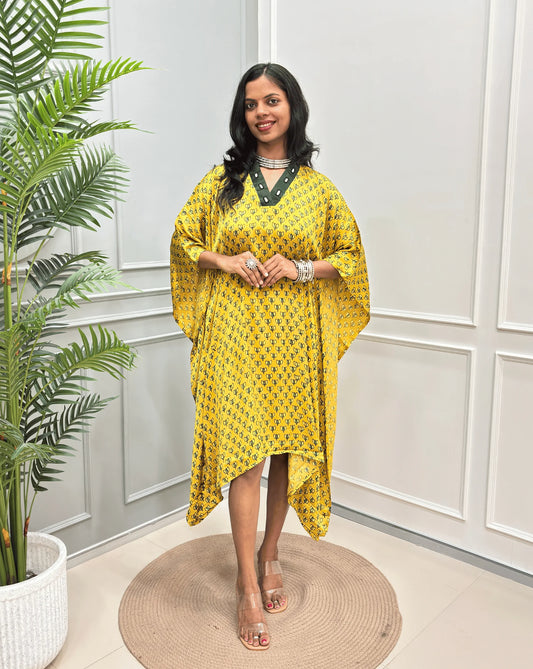 Yellow Women's Ajrakh Pure Modal Silk Knee Length Kaftan with a black Floral print , standing on a round woven rug in front of a white paneled wall next to a potted plant.