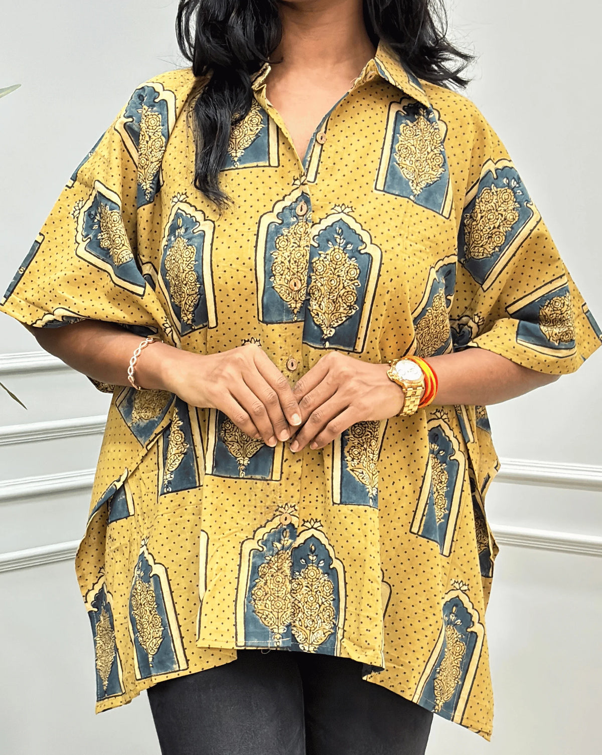 Close-up view of Light Yellow Women's Ajrakh Pure Cotton Short Kaftan Shirt Featured with Shirt Collar neck with a Beige, Green & Yellow Floral Block Print, Wooden Button on front, paired with beige flared pants, standing on a round woven rug in front of a white paneled wall with a potted plant.