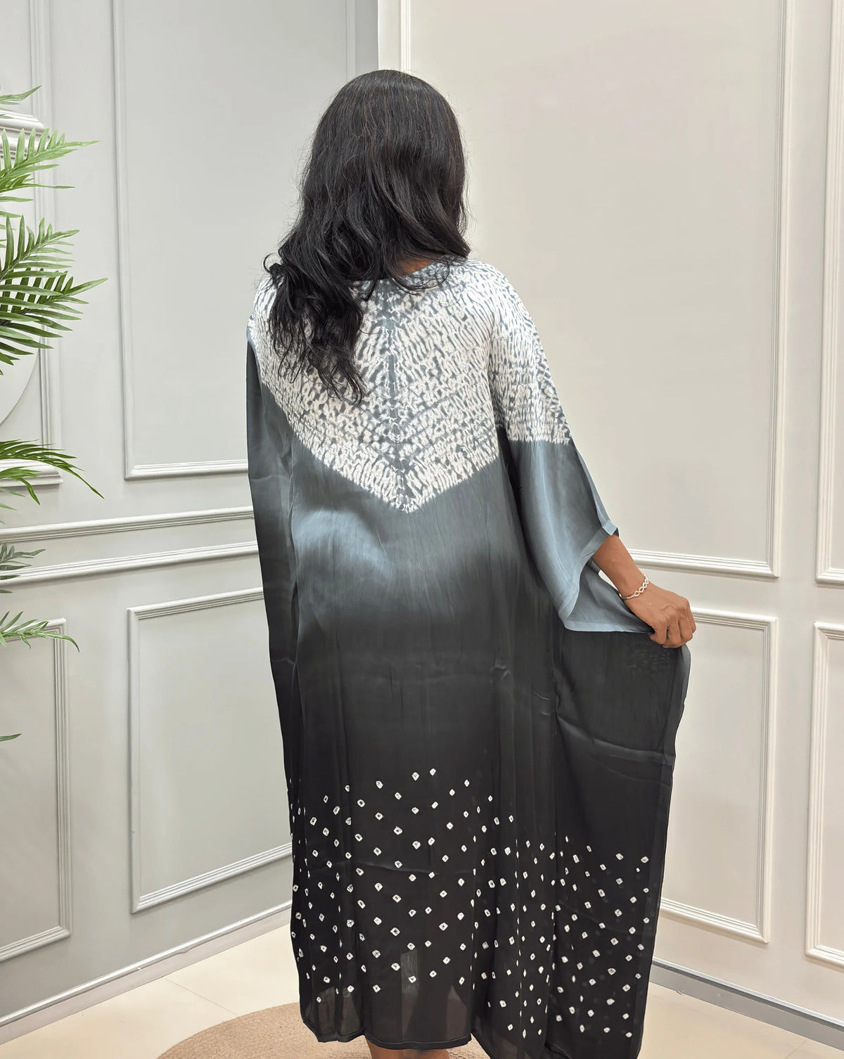 Back-Side View of Grey Women's Ajrakh Pure Modal Silk Shibori Kaftan Featured with Long-length, with a 3 shades of Grey with ajrakh print on chest & Bandhani Small dots with white on Bottom, standing on a round woven rug in front of a white paneled wall.