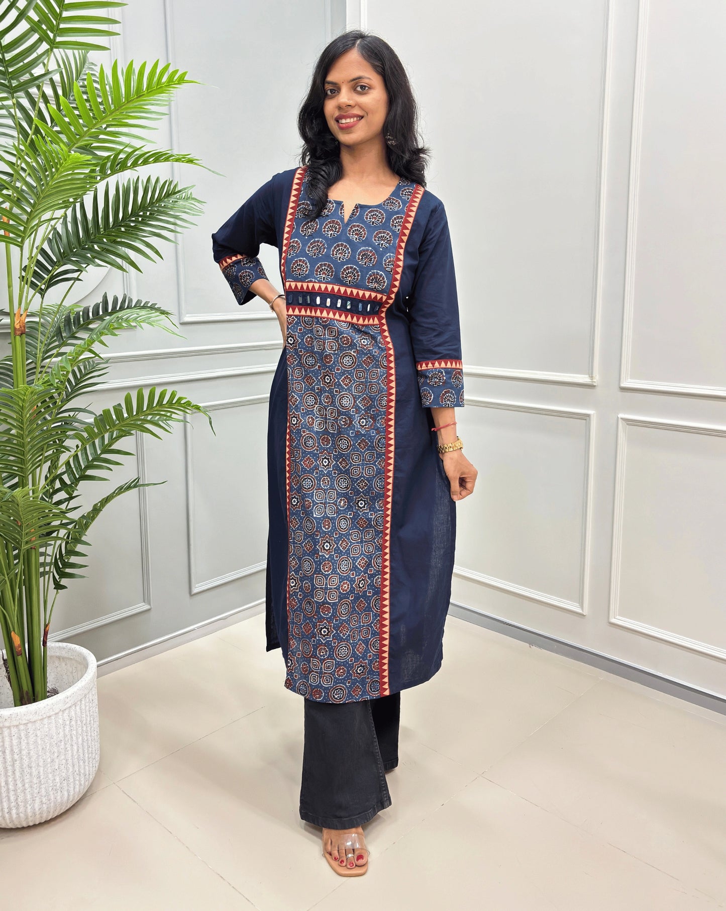 Navy Blue women's Ajrakh Hand-block vegetable dyed Cotton Long-kurti Featuring red and beige border details on font and sleeves, paired with black pants, standing in a bright room with a potted plant on the left.