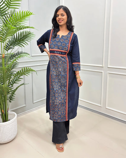 Navy Blue women's Ajrakh Hand-block vegetable dyed Cotton Long-kurti Featuring red and beige border details on font and sleeves, paired with black pants, standing in a bright room with a potted plant on the left.