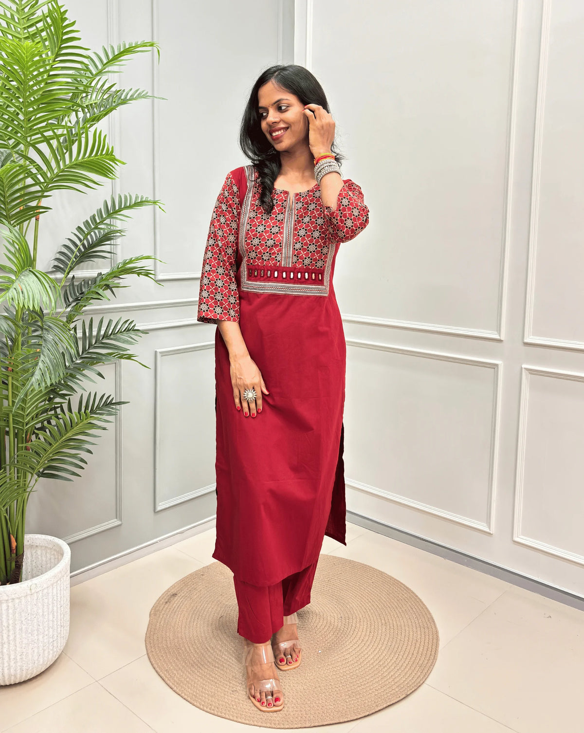 Dark Red Women's Hand-block Ajrakh Pure Cotton Long Kurti with a Red base , an abstract of black & beige Floral Print yoke on front & Full sleeves, paired with blue Pant, standing on a round woven mat in front of a white paneled wall.