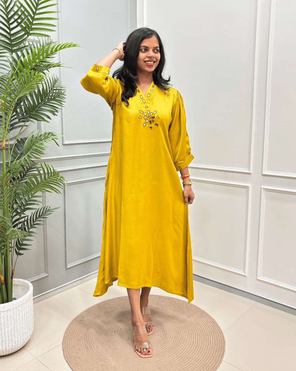 Yellow Women's Pure Modal Silk Ajrakh Long Kurti with a decorative Real Hand-Mirror embroidered neckline, Floral pattern on front & Sleeves Cuffs, standing on a round woven rug in front of a white paneled wall with a potted plant on the left