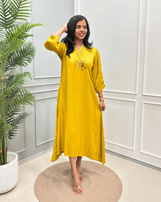 Yellow Women's Pure Modal Silk Ajrakh Long Kurti with a decorative Real Hand-Mirror embroidered neckline, Floral pattern on front & Sleeves Cuffs, standing on a round woven rug in front of a white paneled wall with a potted plant on the left