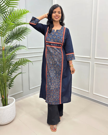 Navy Blue women's Ajrakh Hand-block Cotton long-kurti with intricate blue and red geometric patterns, Hand-mirror work on front like a belt, paired with black wide-leg pants, standing in a bright room with a potted plant on the left.