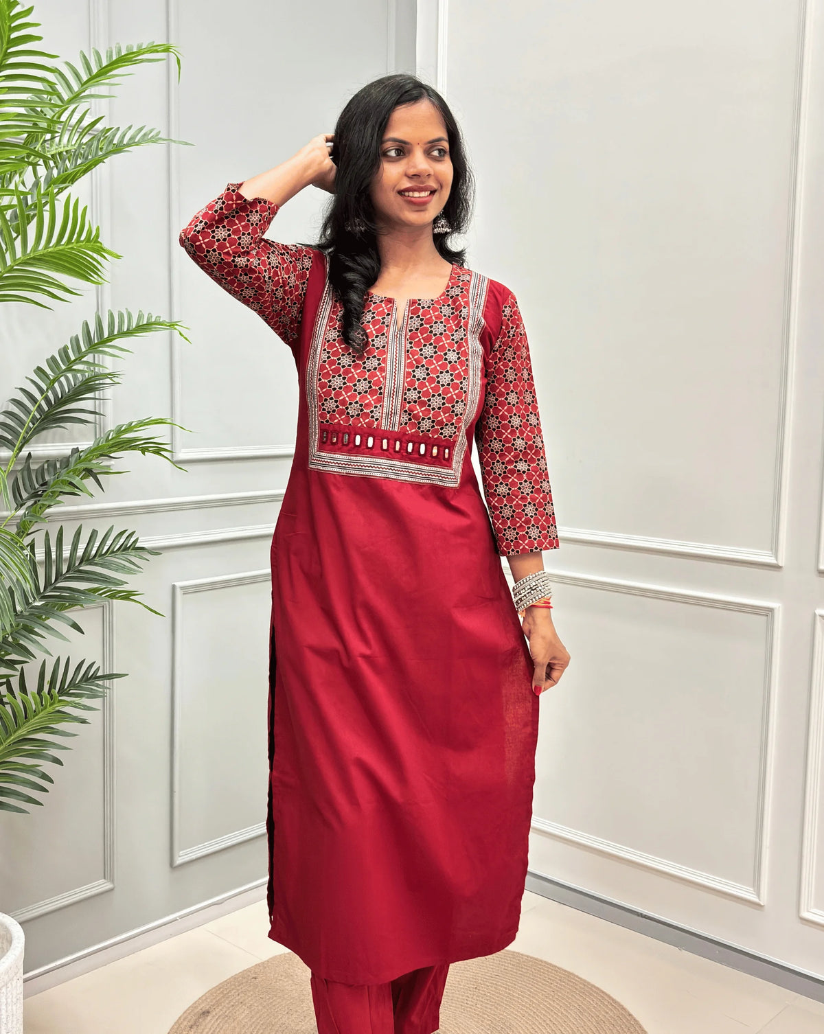 Dark Red Women's Hand-block Ajrakh Pure Cotton Long Kurti with a Red base , an abstract of black & beige Floral Print yoke on front & Full sleeves, Hand-mirror work on front, paired with blue Pant, standing on a round woven mat in front of a white paneled wall.