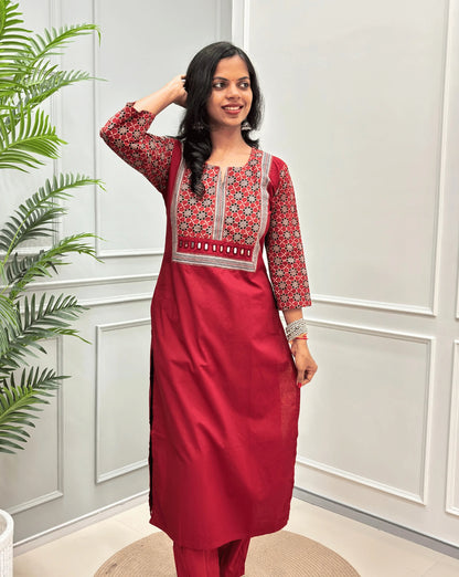 Dark Red Women's Hand-block Ajrakh Pure Cotton Long Kurti with a Red base , an abstract of black & beige Floral Print yoke on front & Full sleeves, Hand-mirror work on front, paired with blue Pant, standing on a round woven mat in front of a white paneled wall.