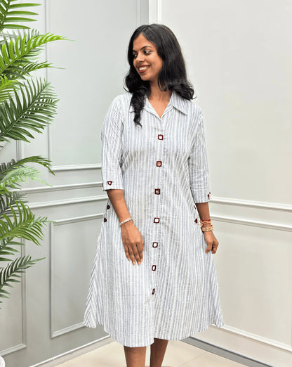 Front-View of Grey Women's Ajrakh A-line Pure Cotton Long Kurti with an Ajrakh Strip Print on abstract of Grey, White,  Real hand-mirror work on front & Sleeves, Along with 2 side pockets, standing on a round woven rug in front of a light gray paneled wall with a potted plant.