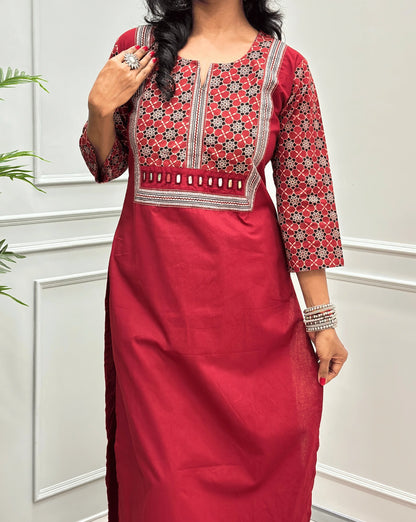 Close-up of Dark Red Women's Hand-block Ajrakh Pure Cotton Long Kurti  featured with Round V Shaped neck along with three quarter sleeves, with a Red base , an abstract of black & beige Floral Print yoke on front & Full sleeves, Hand-mirror work on front, paired with blue Pant, standing on a round woven mat in front of a white paneled wall.