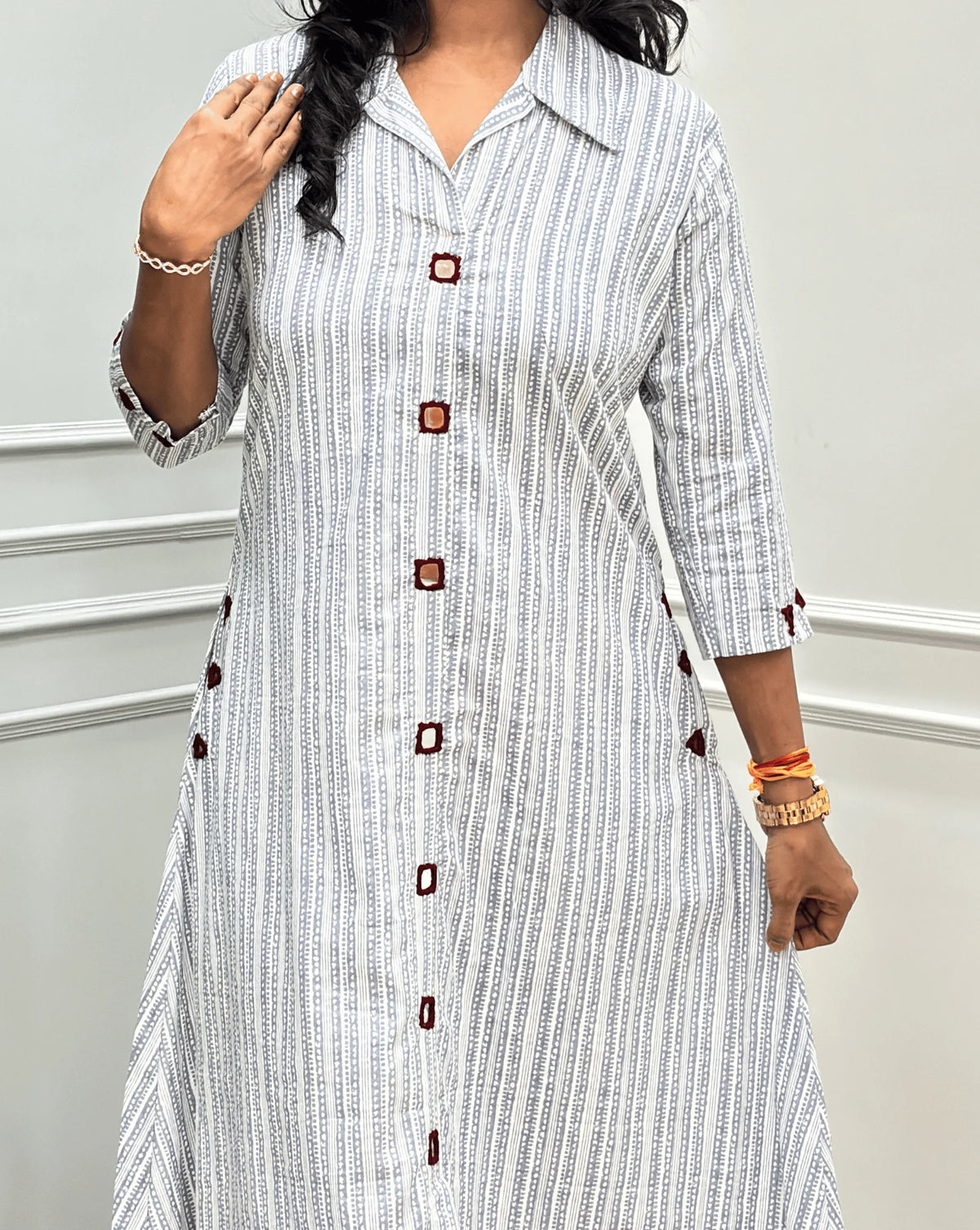 Close-up View of Grey Women's Ajrakh A-line Pure Cotton Long Kurti Featured with Collar V neck & 3/4th Quarter Sleeves with an Ajrakh Strip Print on abstract of Grey, White, Real hand-mirror work on front & Sleeves, Along with 2 side pockets, standing on a round woven rug in front of a light gray paneled wall with a potted plant.