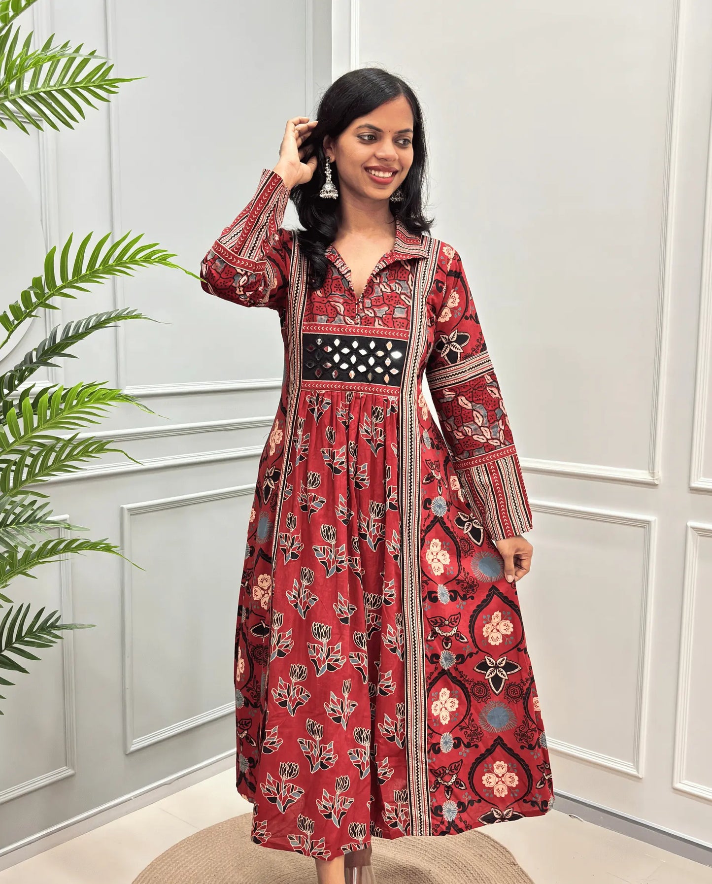  Red women's cotton Ajrakh long kurti front view with mirror work & intricate with floral and geometric patterns, standing in front of a white paneled wall with a green plant on the left.