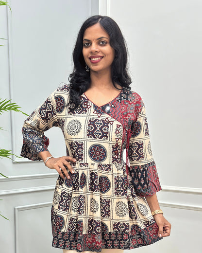Close-up of Beige women's Ajrakh Plant based cotton short-kurti with a Three-Quarter Sleeves (frill)  featuring Red & Black Geometric & block print ,  Angrakha Pattern with Hand-mirror work on V-shaped Neckline along with Lace, set against a light gray paneled wall.